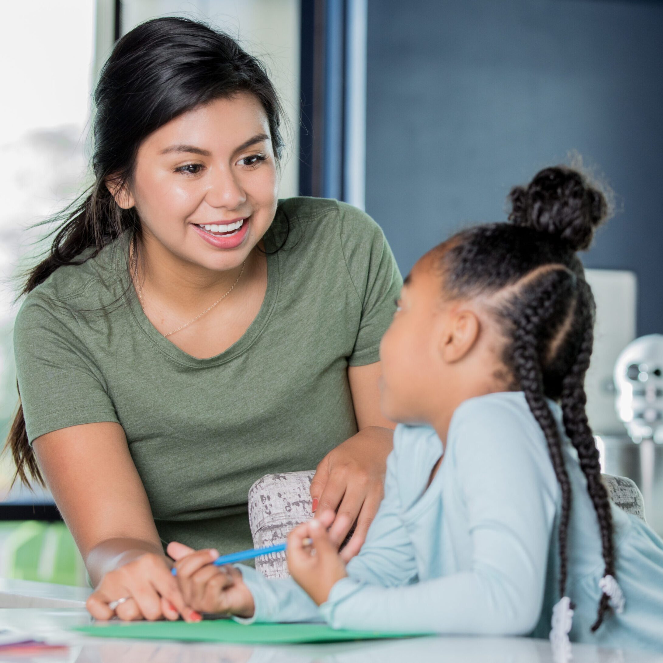 Happy young Hispanic woman is babysitting, tutoring elementary age girl. Young adult Hispanic woman is smiling while babysitting and tutoring African American little girl. Nanny is assisting child with her homework in modern kitchen.