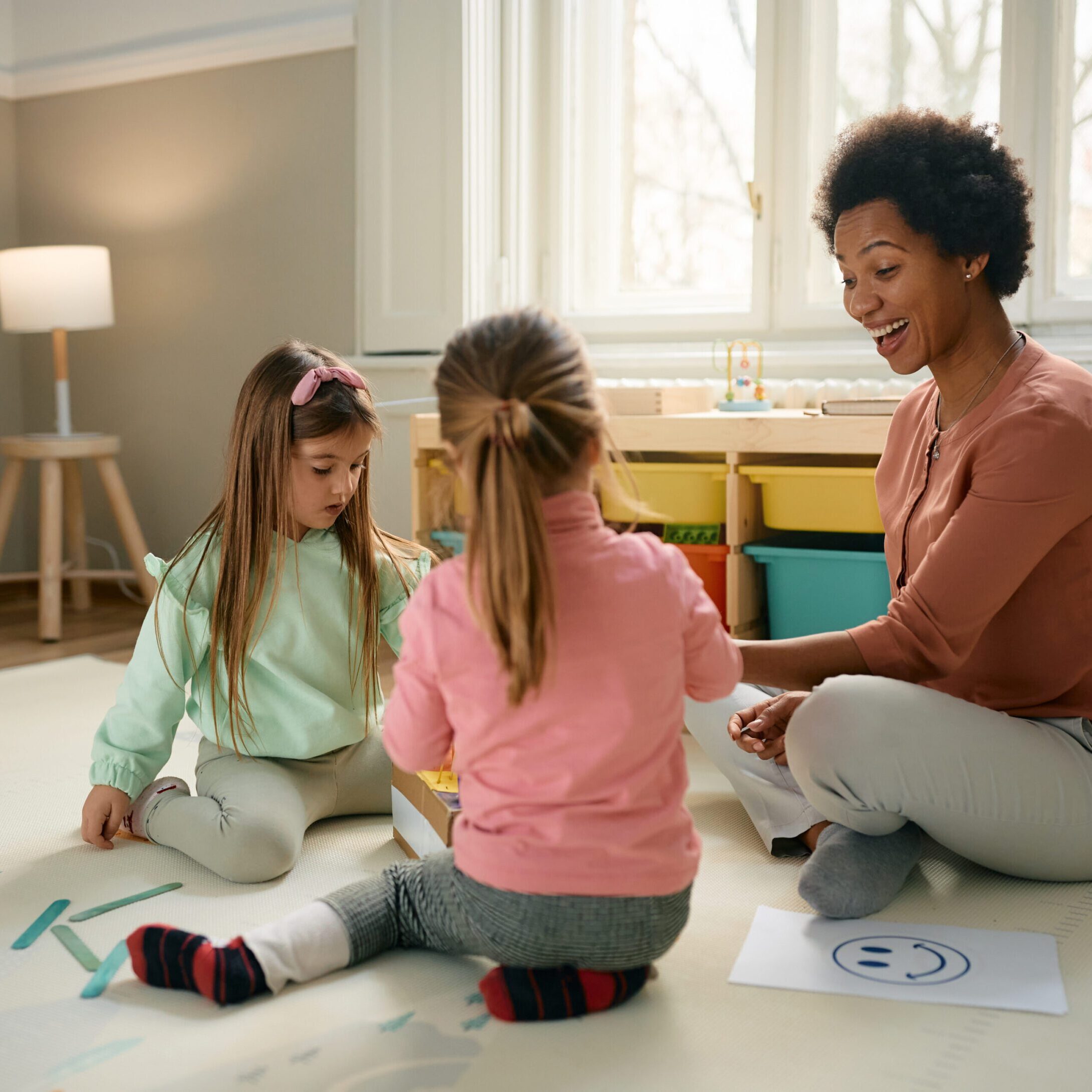 Happy black teacher and small girls having fun while playing at preschool. Happy African American preschool teacher and little girls having fun while sitting on the floor and playing.